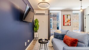 Una sala de estar moderna con un sofá gris, un televisor de pantalla plana montado en la pared, una planta en maceta y un ventanal con vista a un edificio de ladrillo.