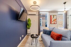 Una sala de estar moderna con un sofá gris, un televisor de pantalla plana montado en la pared, una planta en maceta y un ventanal con vista a un edificio de ladrillo.