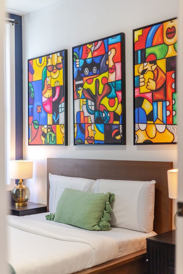A hotel room with a bed, a nightstand, and three colorful abstract paintings on the wall.