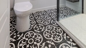 Un baño con inodoro, ducha con mampara de vidrio y suelo estampado.