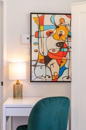 A room with a teal chair, a white desk, a lamp, and a colorful abstract painting on the wall.