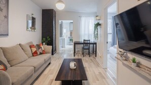Ett modernt vardagsrum med en soffa, ett soffbord och en tv. Det finns en matplats med ett bord och stolar. Rummet har en tegelbeklädd vägg och ett stort fönster.