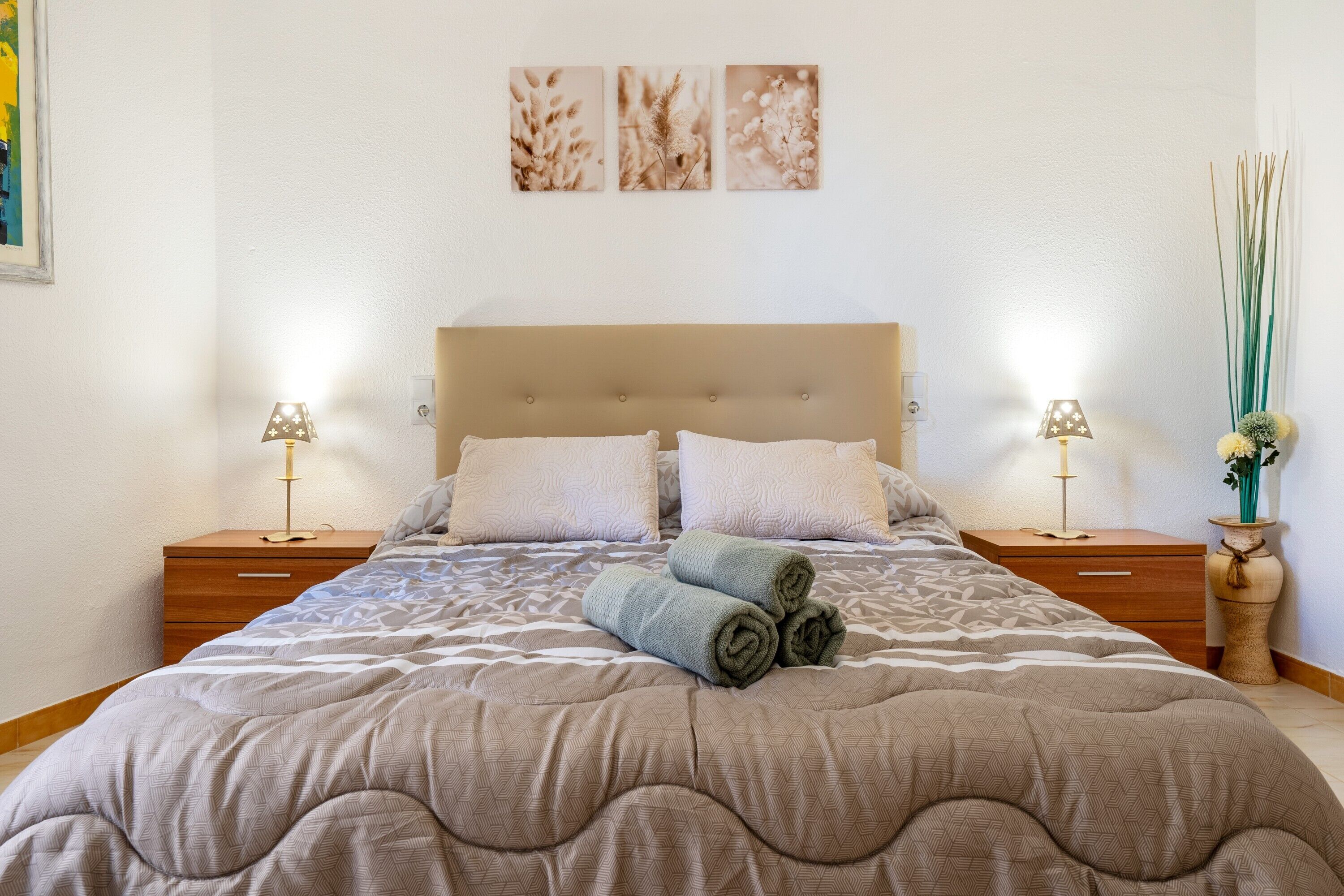 Un lit bien fait, recouvert d’une couvre-lit à motifs, avec deux lampes de chevet et un petit vase rempli de fleurs posé sur une table de chevet.