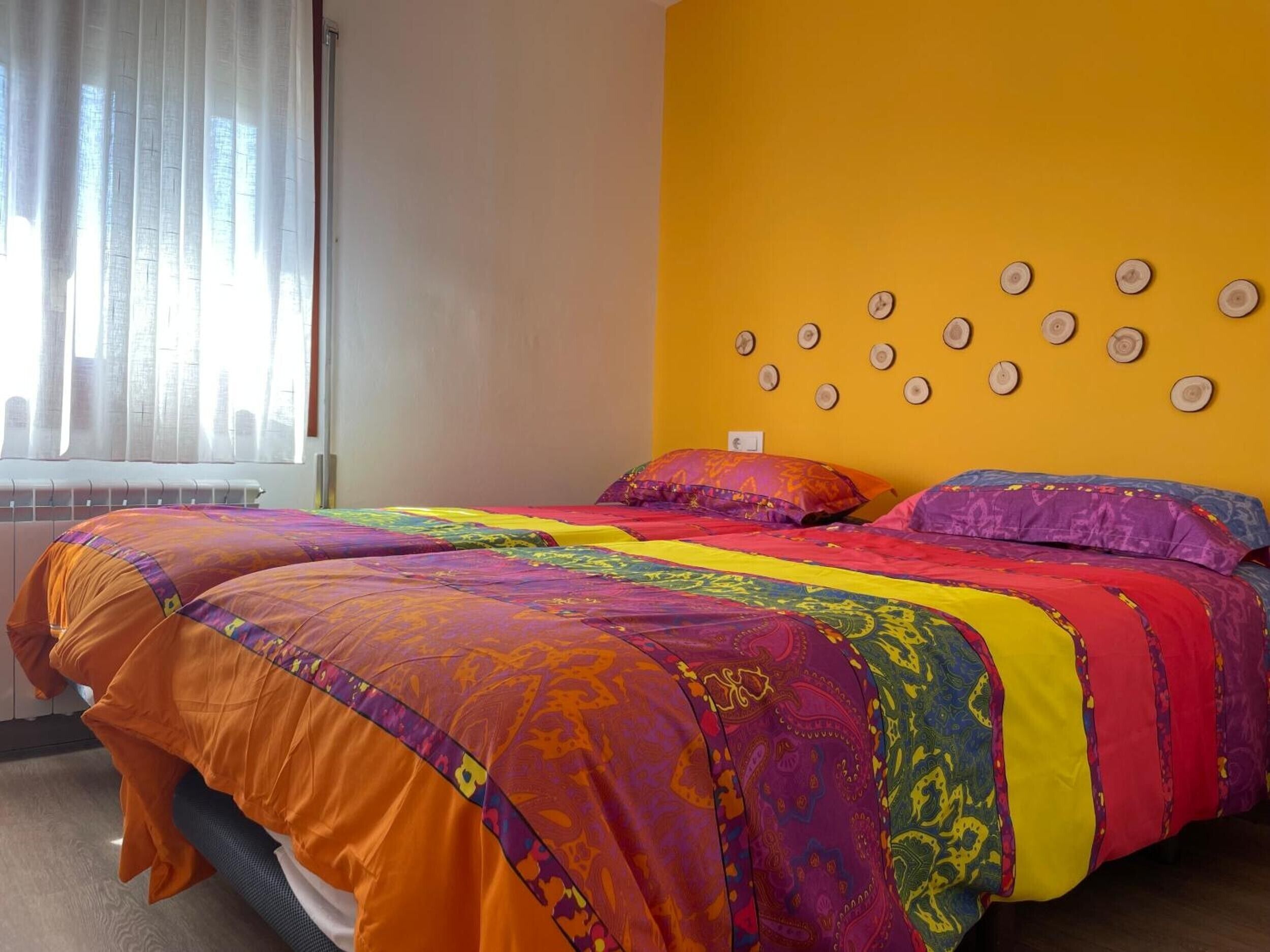 Una habitación con dos camas, una ventana con cortinas traslúcidas, un radiador y una pared con adornos circulares.