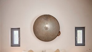 Et soveværelse med en stor seng, to sengeborde med lamper, en rund vægophængning i flet og to små vinduer.
