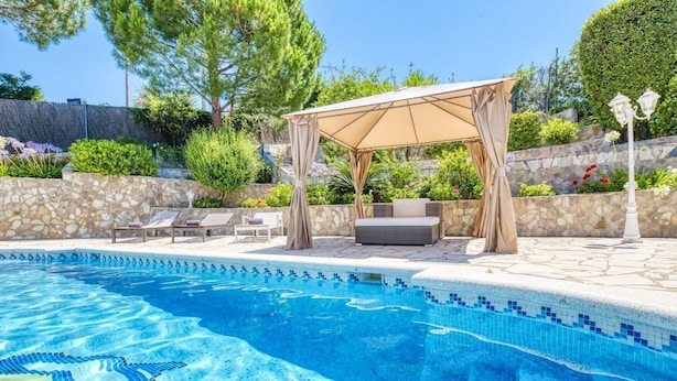 Ein Swimmingpool mit gefliestem Rand, eine überdachte Sitzgruppe mit Kissen und eine Steinmauer im Hintergrund.