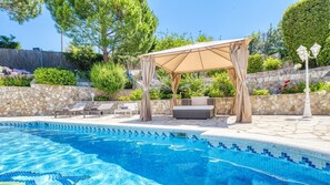 A swimming pool with a tiled edge, a covered seating area with cushions, and a stone wall in the background.