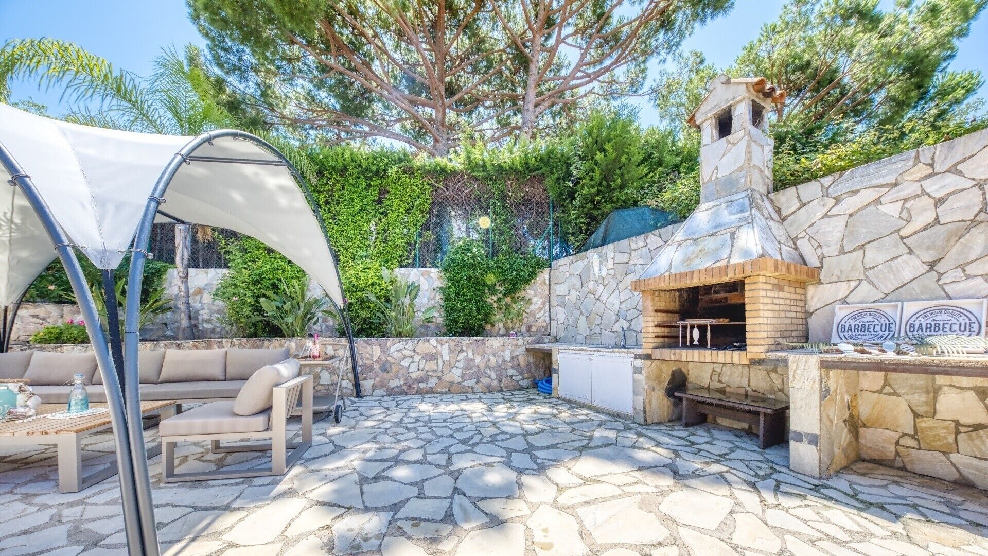 Eine Terrassenanlage im Freien mit Grillplatz aus Stein, überdachter Sitzgruppe und einer Steinmauer mit Bepflanzung.