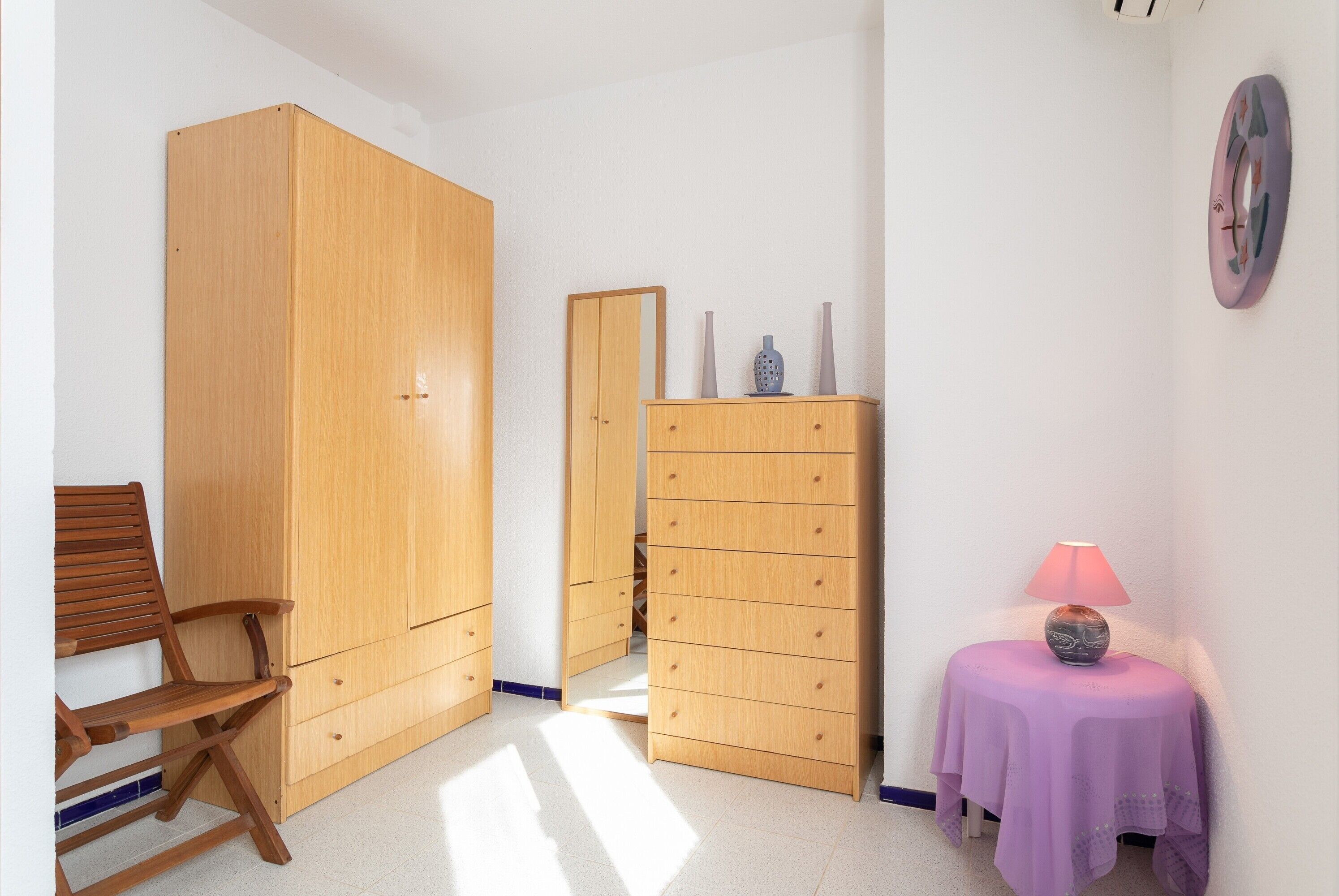 Een kamer met een houten garderobe, een houten stoel, een klein tafeltje met een lamp en een wandklok.