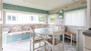 A kitchen with a round table, white chairs, a bench with floral upholstery, and a window with curtains. - Mobile Home for 3 People at Lake Isachris – Hiking and Relaxation in the Médoc (Queyrac)