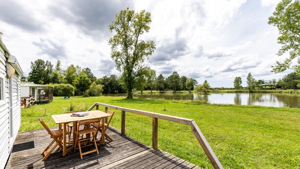 A wooden deck with a table and chairs, overlooking a lake and grassy area. - Mobile Home for 3 People at Lake Isachris – Hiking and Relaxation in the Médoc (Queyrac)