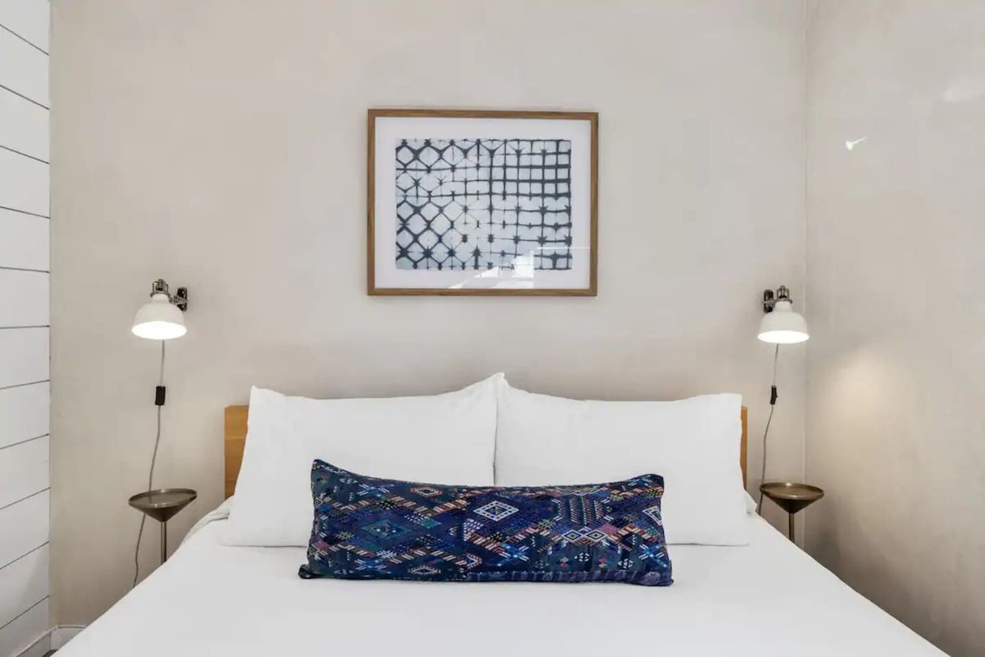 A neatly arranged hotel room with a large bed, two bedside lamps, a framed abstract artwork above the bed, and a patterned throw pillow.