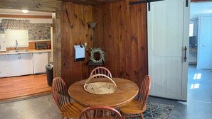 Zona pranzo con un tavolo rotondo in legno, quattro sedie di legno, un sottopiattino intrecciato, una lampada a parete e una ghirlanda decorativa.