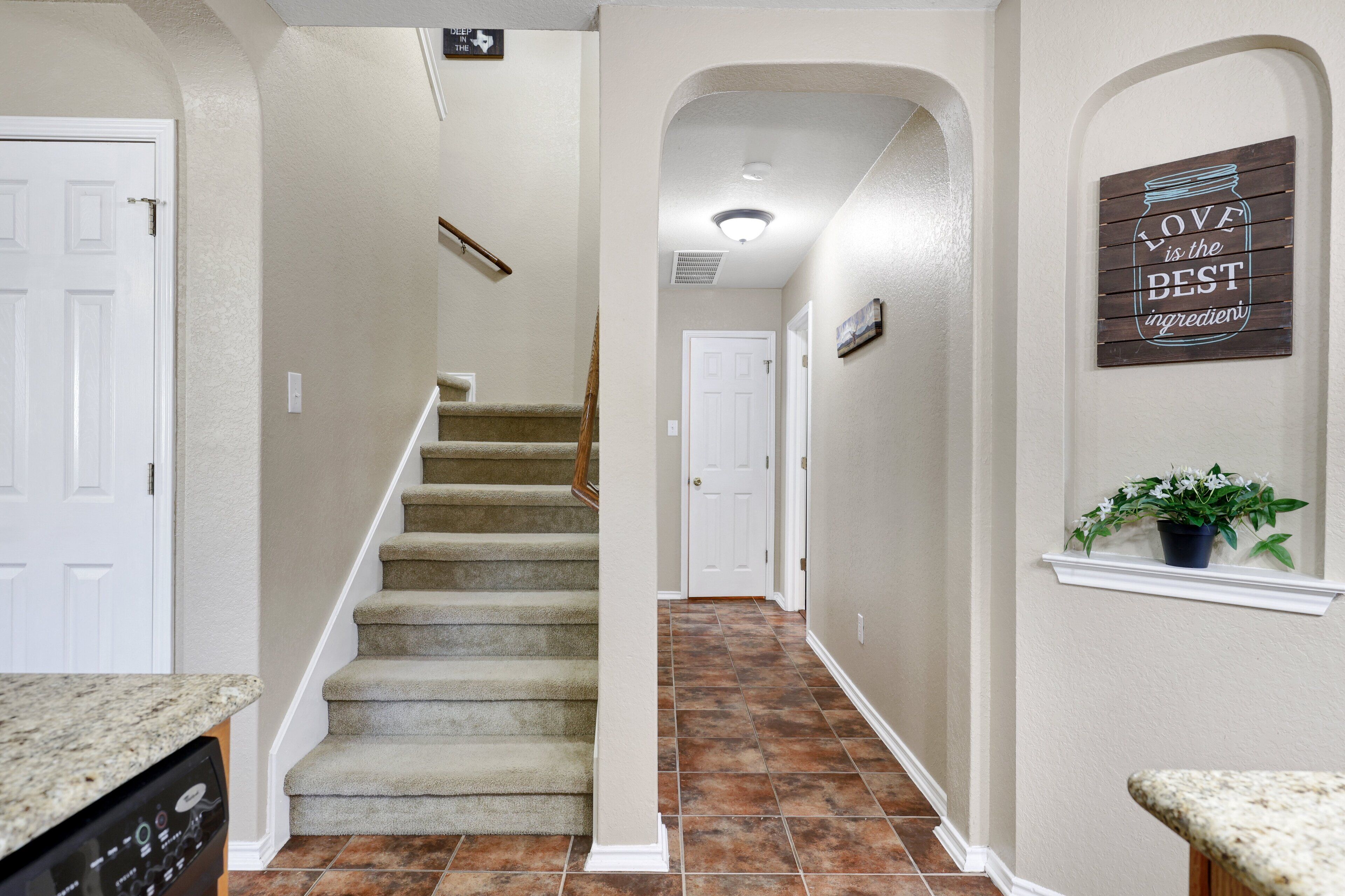 A hallway with a carpeted staircase, a white door at the end, and a wall-mounted sign that says "LOVE is the BEST ingredient."