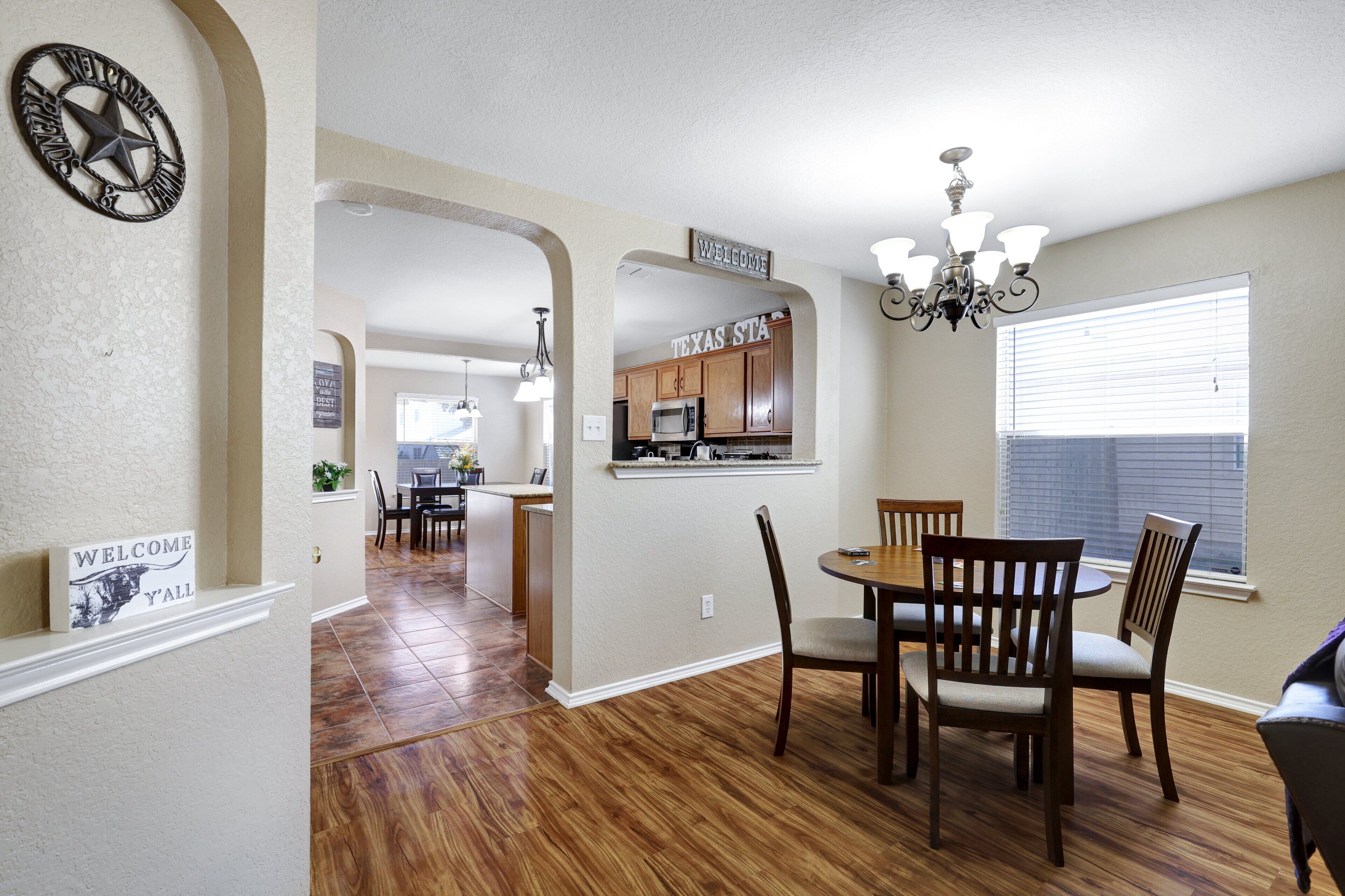 A spacious dining area with a wooden table and chairs, a kitchen in the background, and a chandelier.