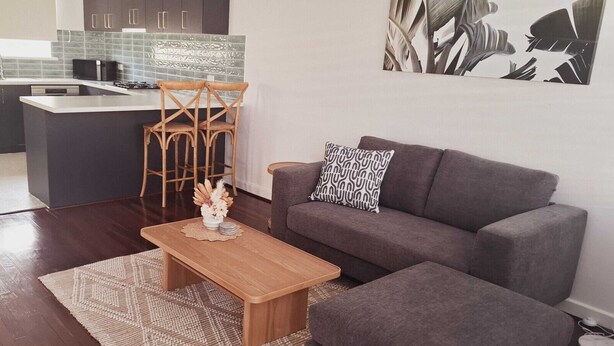 A modern living room with a grey sofa, a wooden coffee table, and a kitchen area with dark cabinets and a microwave. - Beachside Homely Flat in Rockingham (Rockingham)