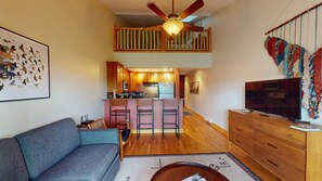 A living room with a sofa, a TV, a ceiling fan, and a kitchen area with a bar. - Luxury Studio | In Town | Wi-Fi | The Grande 312 (Angel Fire)