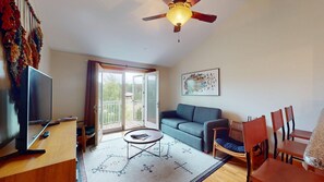 A living room with a television, a sofa, a dining table with chairs, a ceiling fan, and a sliding glass door leading to a balcony.