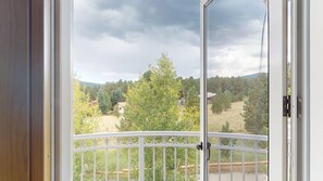 A view through a balcony door with a white metal railing, overlooking a landscape with trees and buildings.