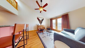 Ein modernes Wohnzimmer mit Deckenventilator, einer Couch, zwei Sesseln, einem kleinen Tisch und Blick ins Freie durch ein großes Fenster.