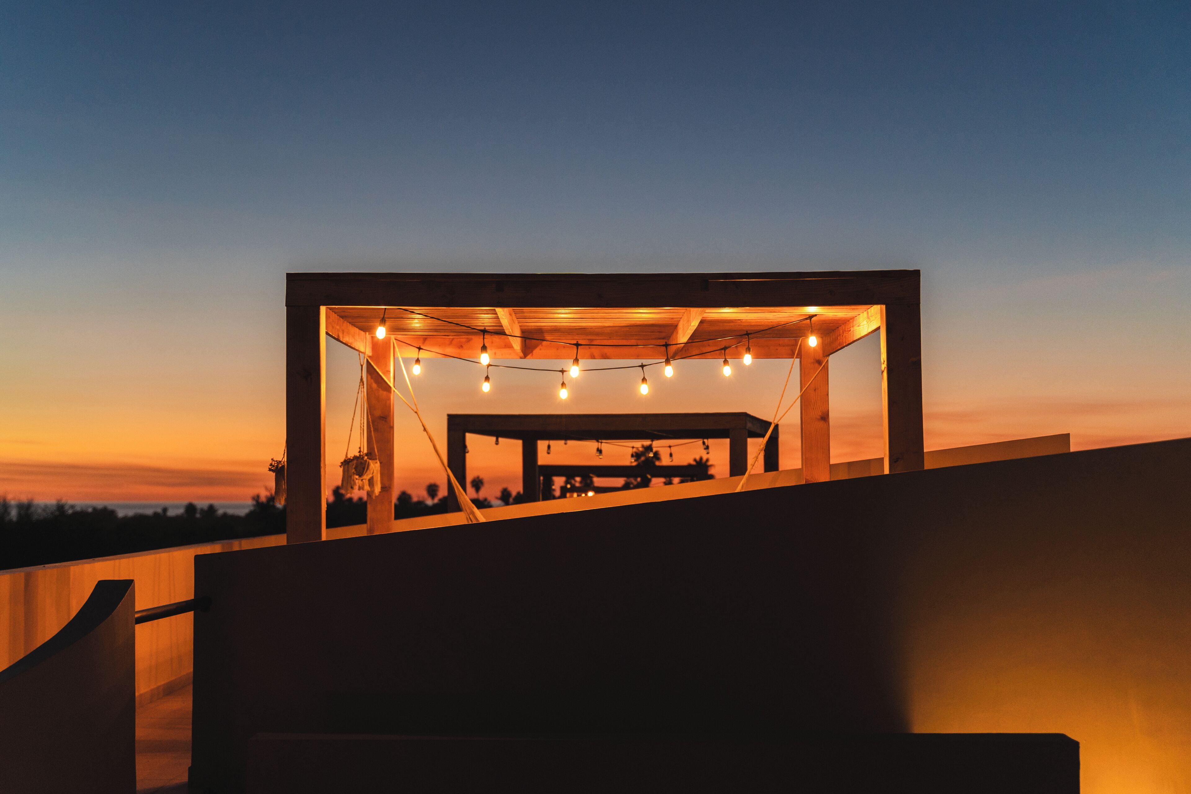 A modern outdoor seating area with string lights, a wooden structure, and a view of the sunset.
