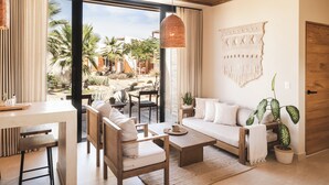 Ein modernes Hotelzimmer mit einem großen Bett, einer Sitzecke mit Sofa und Sesseln, einem Esstisch und Blick auf einen Garten mit Palmen.