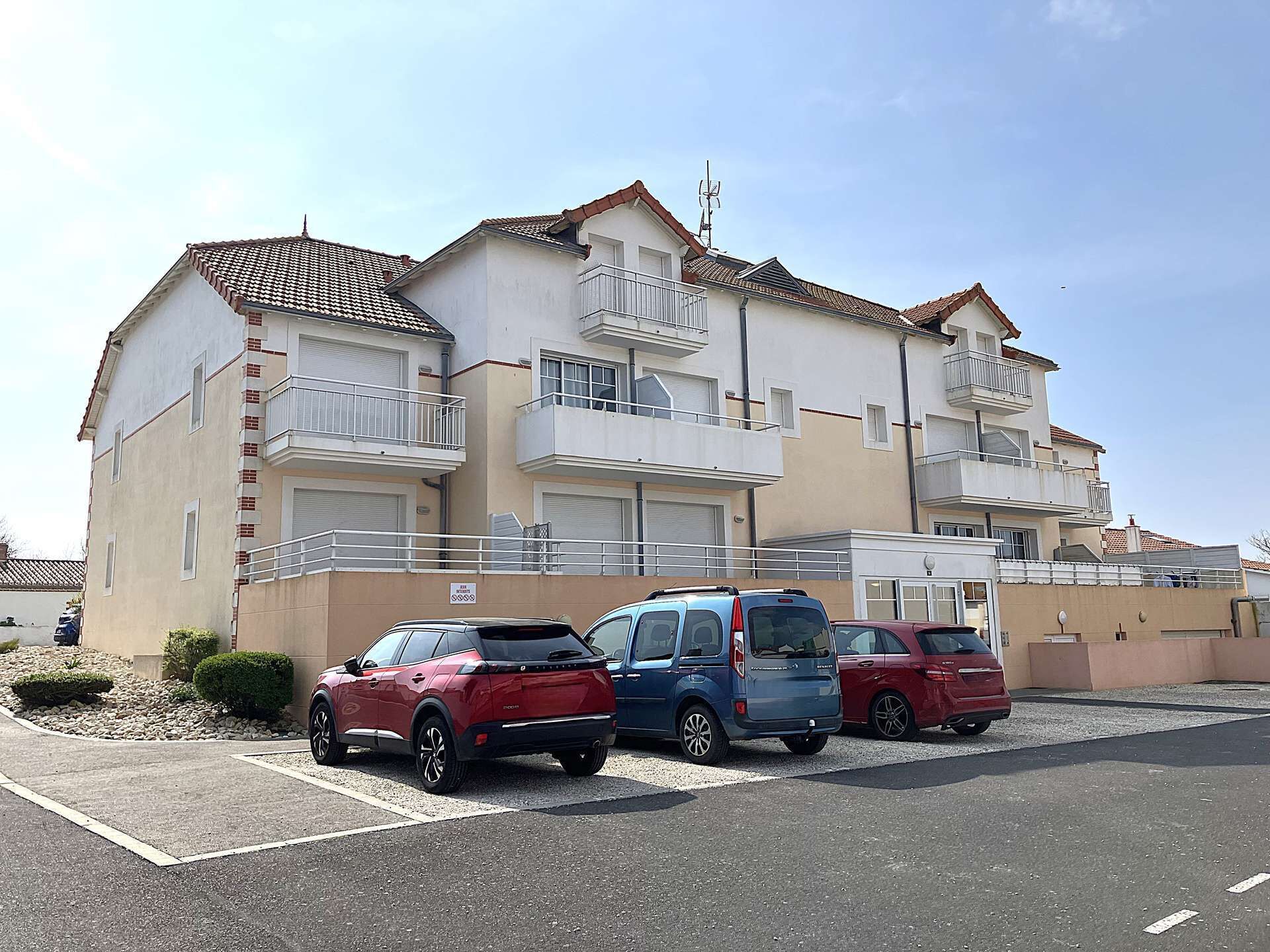 Un edificio residencial de varios pisos con balcones, un estacionamiento y tres coches aparcados.