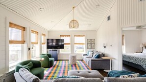 A modern living room with a flat-screen TV, green and grey sofas, a colorful striped rug, and a bed with a patterned duvet. - 1 Block from the Beach, Entertainment Deck with games and Tv, large enough for 26 guests. (Bolivar Peninsula)