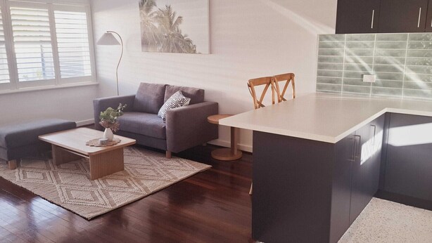 Una sala de estar moderna con un sofá, una mesa de centro y una pequeña mesa de comedor. Hay una zona de cocina con armarios oscuros y un salpicadero de azulejos.