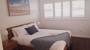 Un dormitorio ordenado con cama, almohadas, lámpara de noche, ventana con persianas y un cuadro con motivos playeros en la pared.