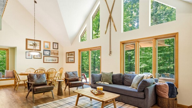 A spacious living room with a sofa, chairs, a coffee table, and large windows offering a view of trees. - Up North Chalet | Ac | Bbq | Bikes (Cedar)