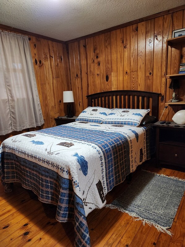 A bedroom with wooden panel walls, a bed with a quilt featuring fish patterns, a nightstand with a lamp, and a small shelf with books. - Riverfront! Snappin Turtle Cabin, Balcony-Floating dock-Fishing. (Steinhatchee)