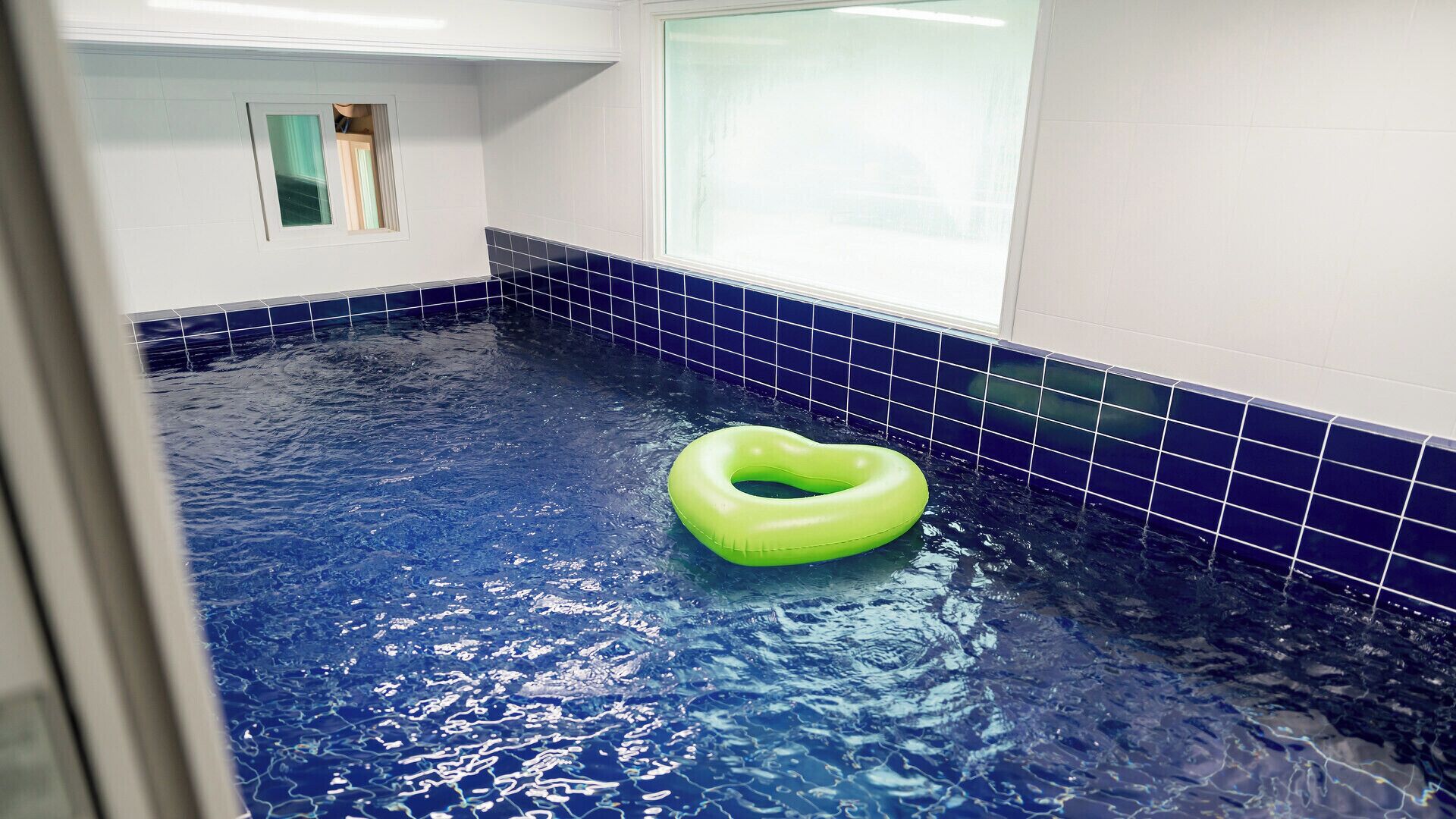Una piscina con un flotador verde, paredes de azulejos y una ventana.