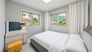 A bedroom with a bed, a desk, a chair, and two windows with a view of greenery. - Gapyeong Dream Pool Villa Pension (Gapyeong)