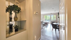 A modern interior with a hallway leading to a bedroom and a dining area. There are potted plants on a glass shelf, a bed with a nightstand, and a dining table with chairs.