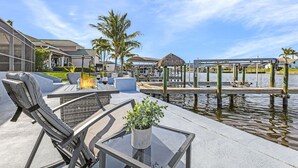 An outdoor patio area with a glass table, cushioned chairs, a small fire feature, and a water view. - Direct Gulf Access, Heated Pool - Sunshine Getaway - Roelens (Cape Coral)