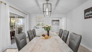 A dining area with a marble table, six chairs, a vase with flowers, and a bottle of wine. - Direct Gulf Access, Heated Pool - Sunshine Getaway - Roelens (Cape Coral)