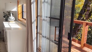 Una habitación con puerta de vidrio corrediza que da a una terraza de madera, un escritorio blanco con lámpara y vistas a árboles.