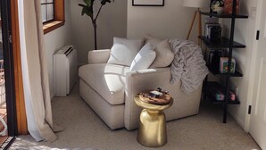 Una sala de estar acogedora con un sofá beige, una mesita auxiliar redonda dorada, una planta verde grande, un cuadro enmarcado en la pared y una estantería negra con libros y una lámpara.