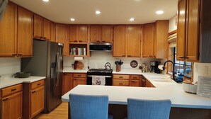 Una cocina moderna con armarios de madera, un refrigerador de acero inoxidable, una estufa y un lavamanos. Hay una zona de bar con dos sillas azules.