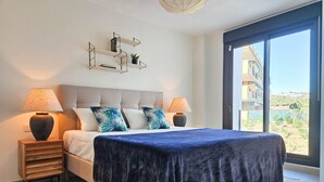 A bedroom with a bed, two bedside tables, a nightstand, a large window, and a view of buildings outside.