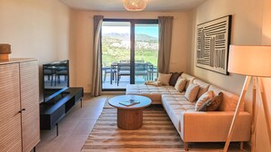 A modern living room with a sofa, coffee table, TV, and a dining area with a view of mountains.