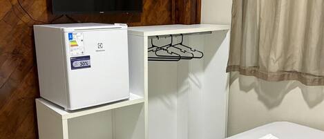 A hotel room with a bed, a white minibar, a television mounted on a wooden wall, and a coat rack.
