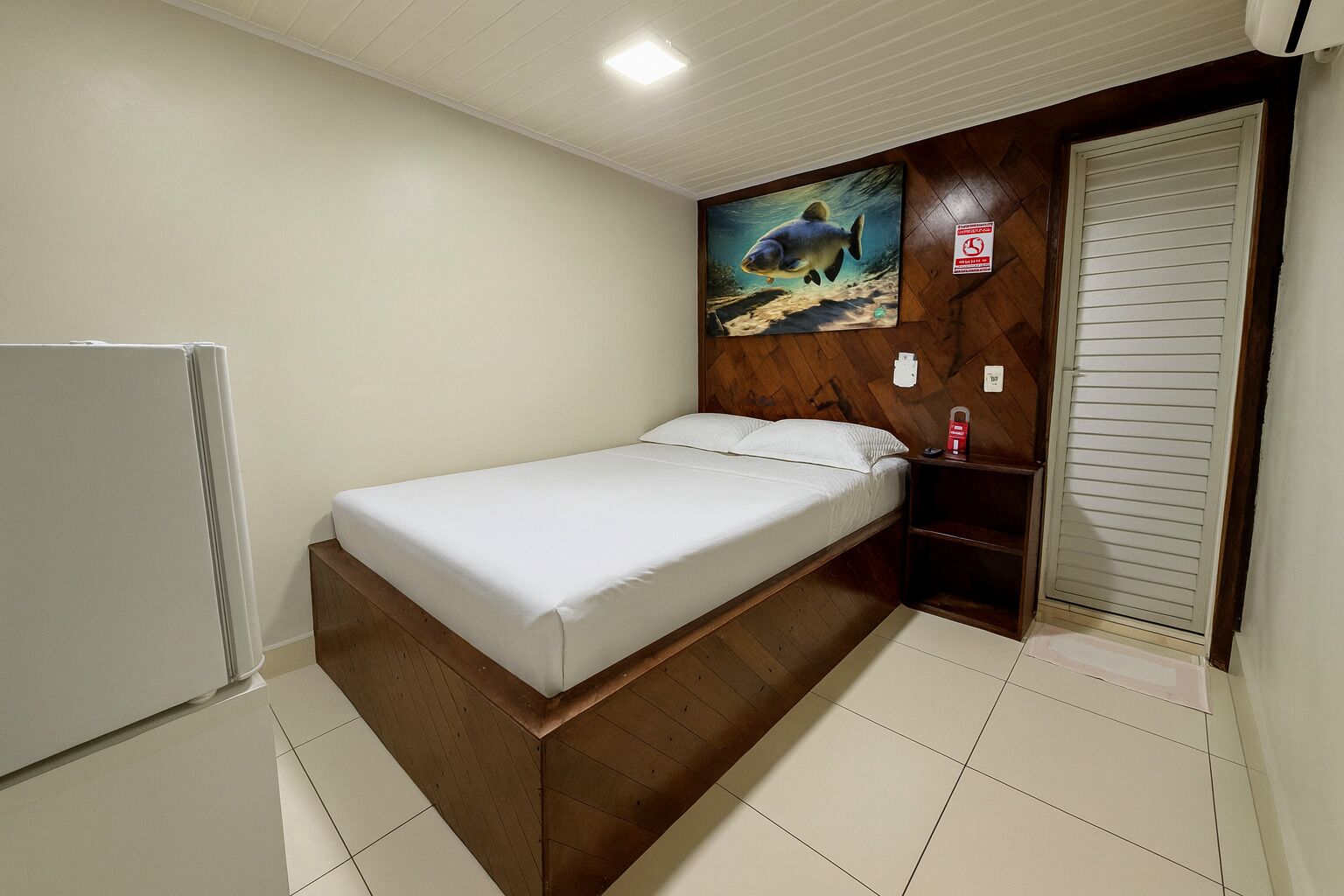 A bedroom with a bed, a wooden headboard, a painting of a fish, a door with a sign, and a white refrigerator.