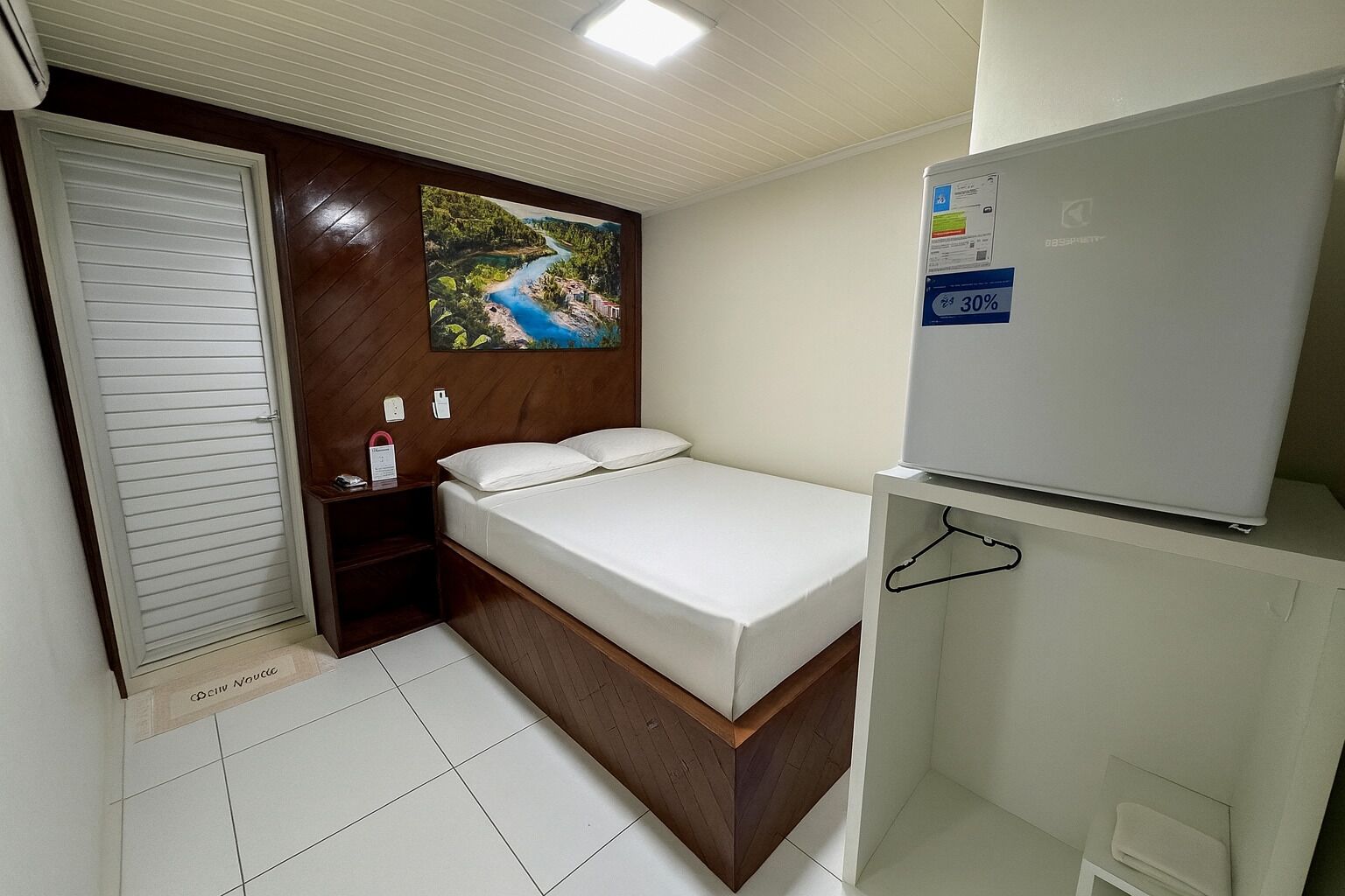 A hotel room with a bed, a wall-mounted TV, a whiteboard, and a painting of a landscape.