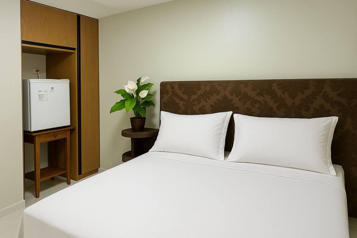 A hotel room with a bed, a wardrobe, a small table with a potted plant, and a refrigerator.