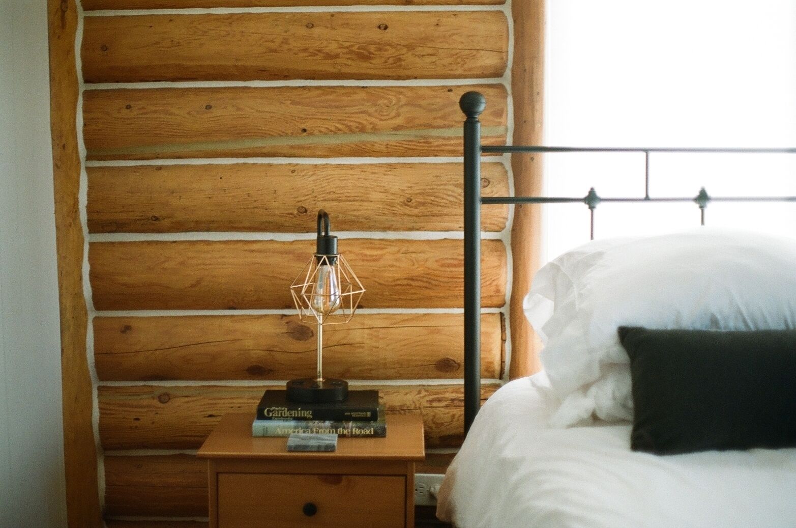 A wooden log wall, a metal bed frame, a bedside table with a lamp, and a book titled 'Gardening'.