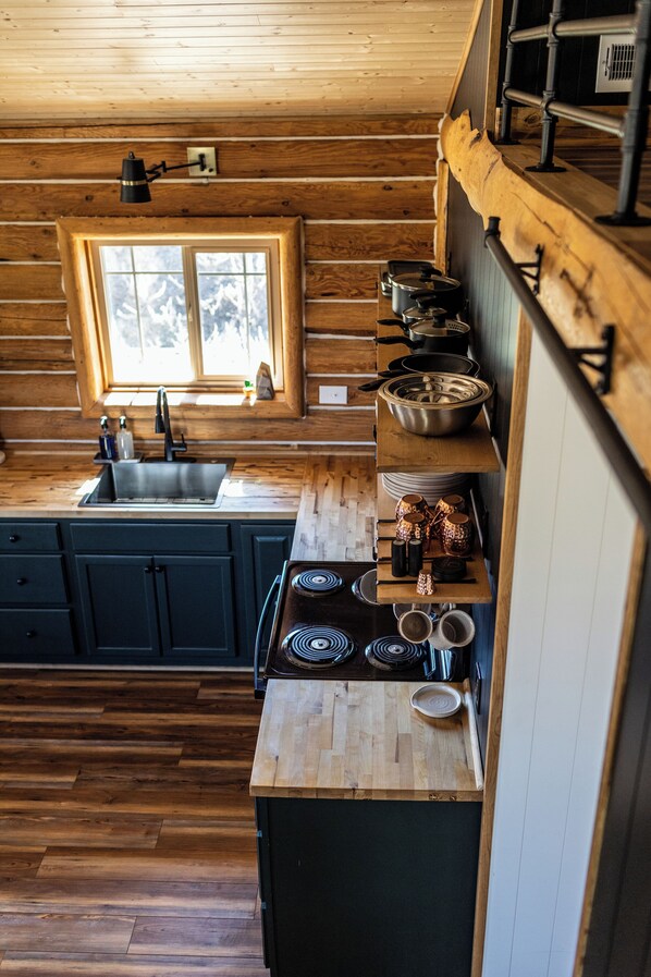 A wooden cabin kitchen with a window, sink, stove, and shelves with cookware.