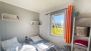 A bedroom with two beds, a window with curtains, a shelf with books, and a plant.
