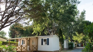 A small, single-story mobile home with a porch and steps, surrounded by trees and a clear path.